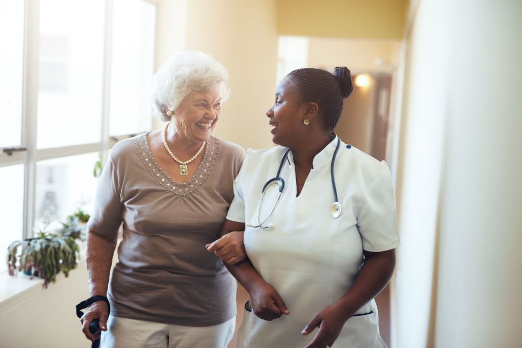 Smiling senior lady walking down hallway with the help of a registered nurse.