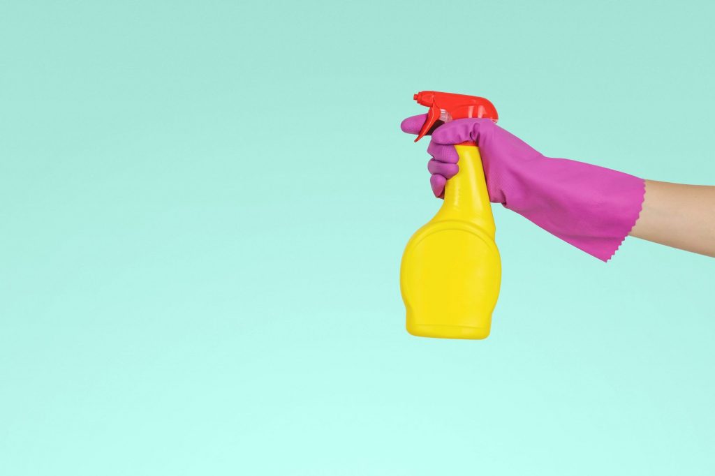 Pink gloved hand brandishing yellow spray bottle in front of mint background.