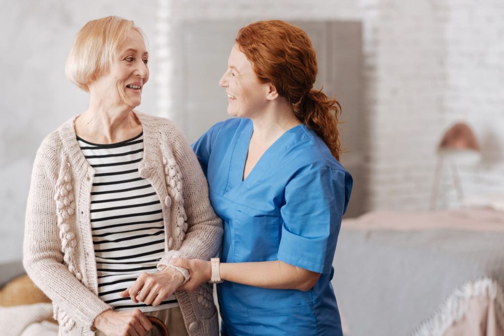 Smiling redheaded nurse helping elderly lady in cardigan. 