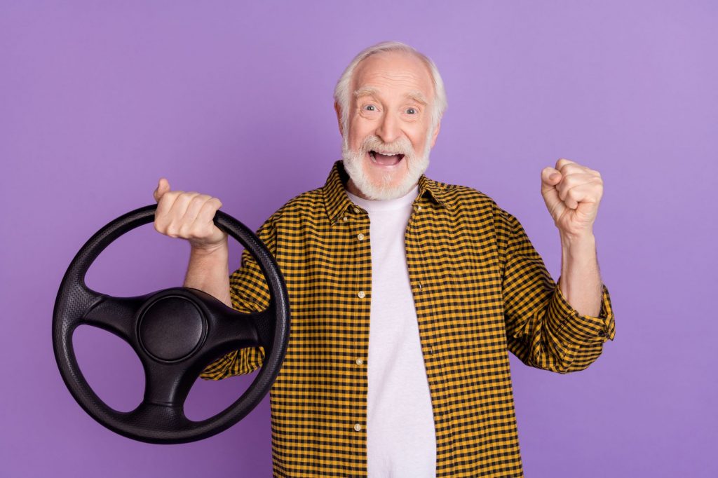 senior man holding a steering wheel against purple background