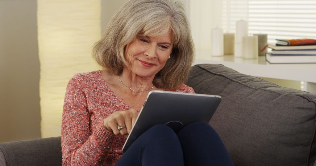 senior woman sitting on couch using a tablet to Track Down Old Friends Online
