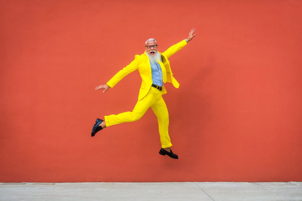 Aging man in yellow suit jumping for joy amid orange backdrop. 