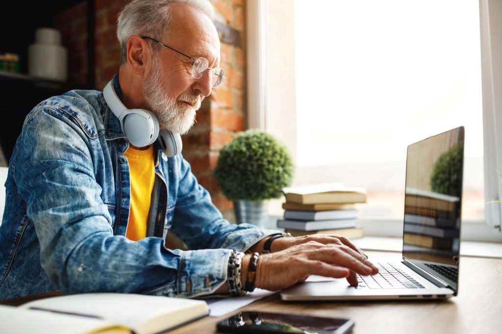 Boomer man wearing a jean jacket and wearing headphones around his neck using a laptop computer at a desk to make money