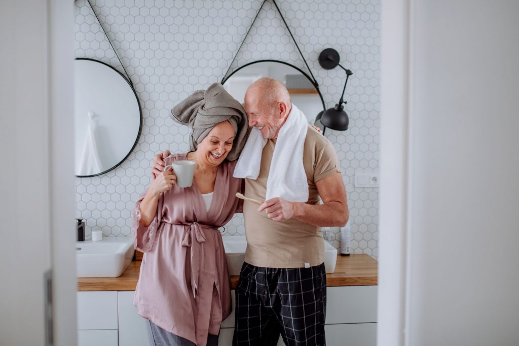 retired couple in bathroom one with coffee cup other with toothbrush