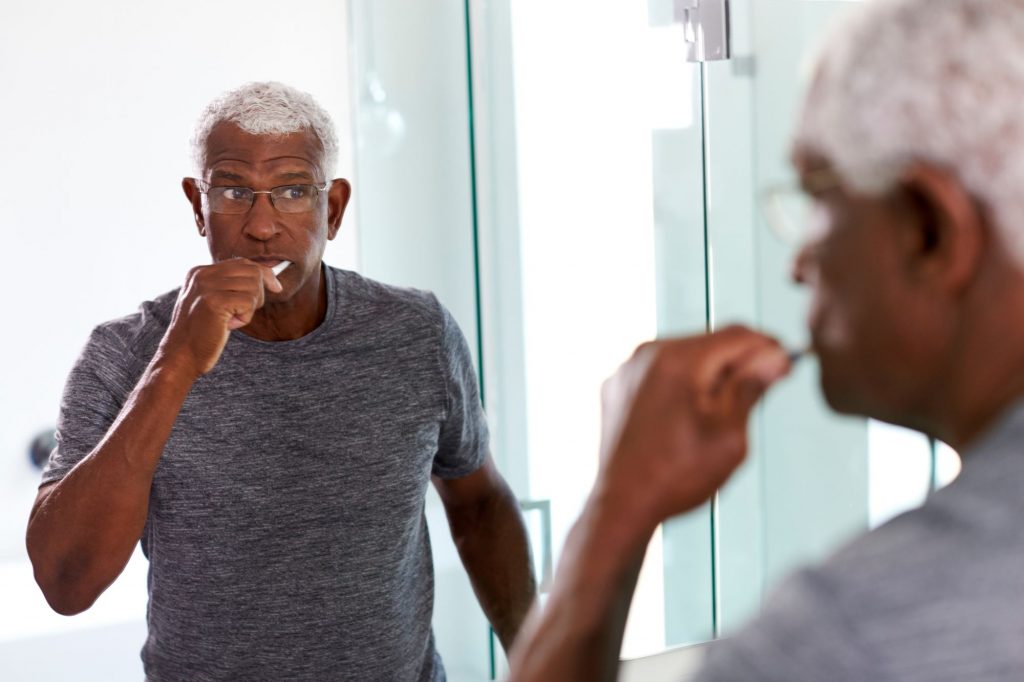 African American retired man brushing his teeth in a mirror, thinking about his dental care