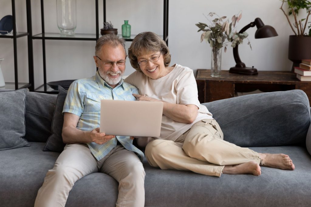retired couple sitting on the couch with a laptop, smiling, planning their retirement abroad
