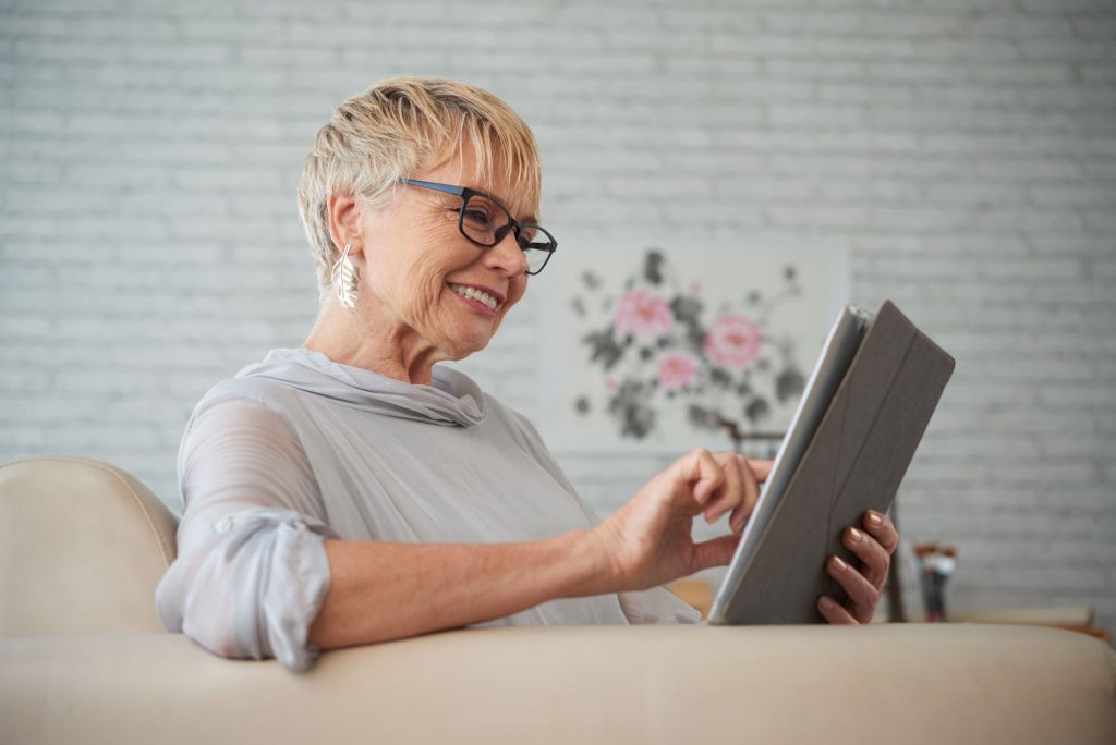 senior woman wearing classes, sitting on couch reading on a tablet