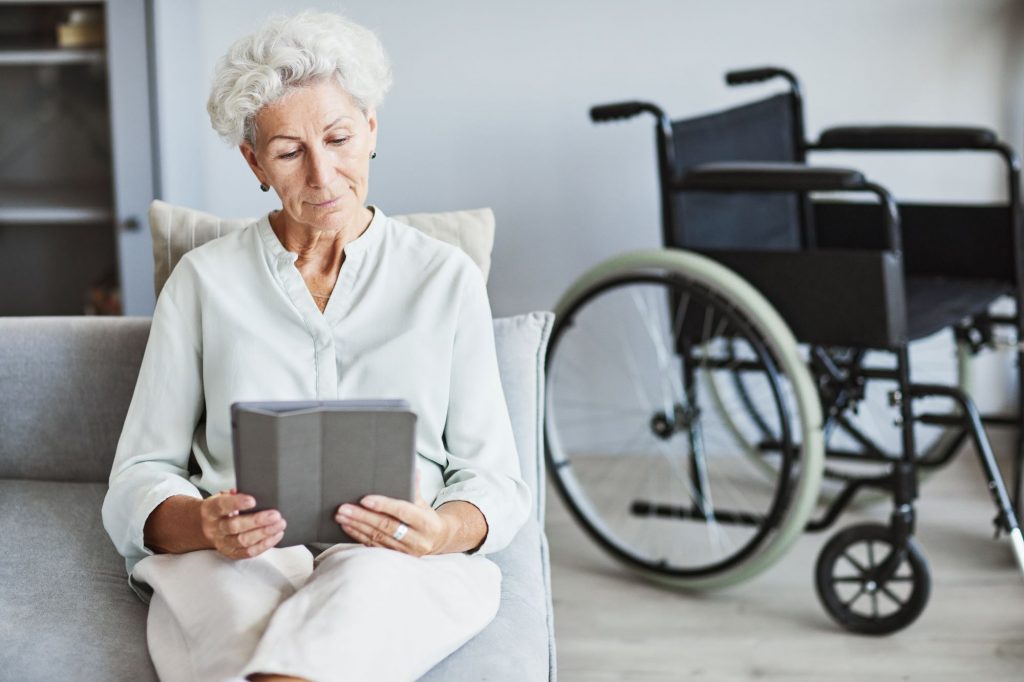 senior woman sitting on the couch reading with her wheelchair parked behind her
