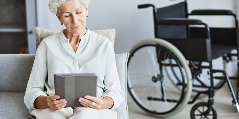 senior woman sitting on the couch reading with her wheelchair parked behind her