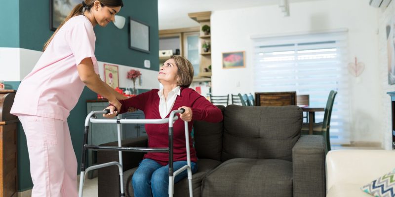 Aide helping woman in long-term care facility stand up with walker