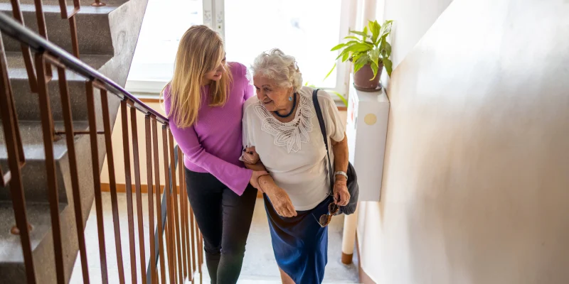 young adult woman helping senior woman up steps in a home