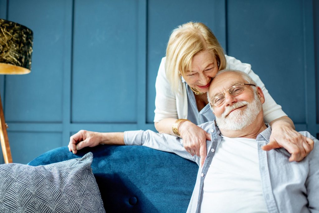 senior couple sitting on a couch, hugging and smiling, with blue paneled walls in the background, symbolizing aging in place