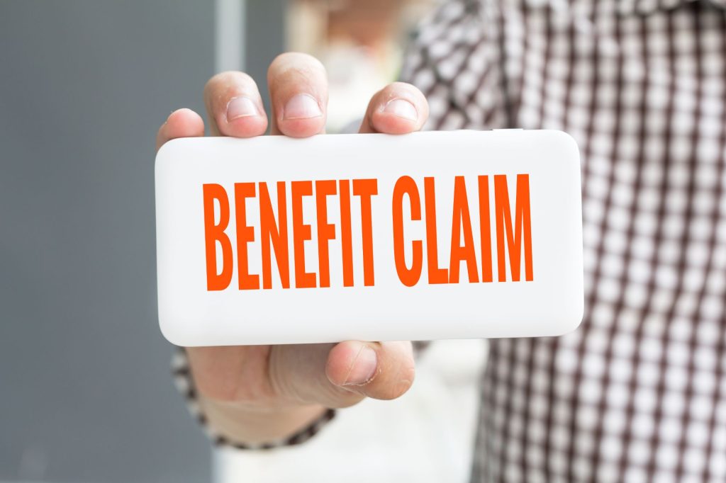 Man hand showing BENEFIT CLAIM word phone with blur business man wearing plaid shirt.