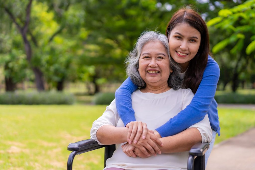 Smiling caregiver in a blue sweater hugging elderly lady in wheelchair. 