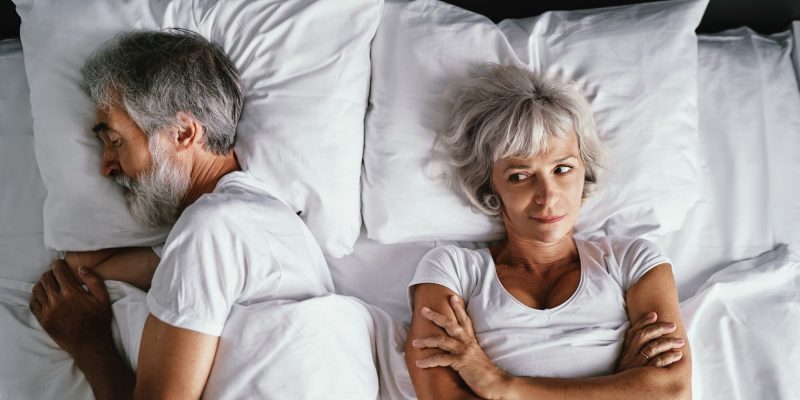 senior couple in bed, white sheets and pillows, man sleeping, woman awake and looking annoyed