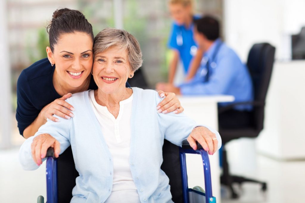 senior woman sitting in a wheelchair, wearing a blue sweater, and smiling with a healthcare worker in a continuing care retirement community
