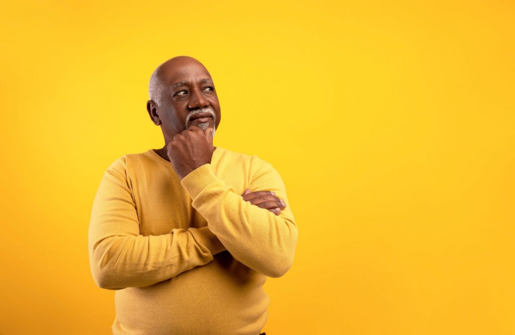black senior man with bald head, wearing a yellow shirt, standing against a yellow background with his arms crossed like he is thinking