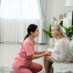 home care aid wearing pink, holding hands of resident on couch
