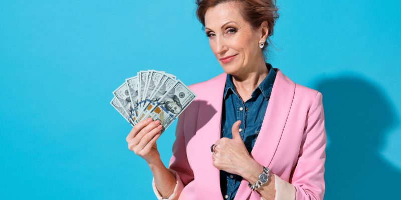 mature older woman with a grin and a fan of hundred dollar bills thumbs up