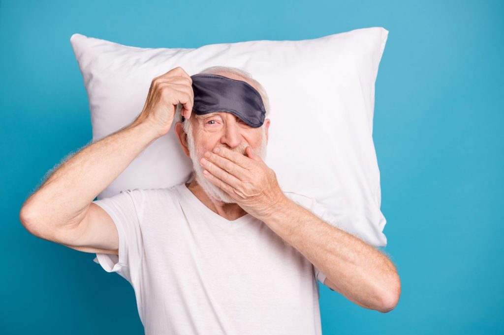 senior with a sleep mask and pillow on a blue background