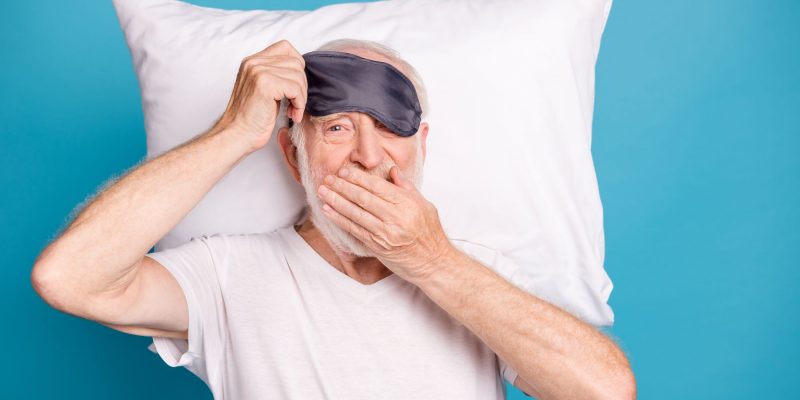 senior with a sleep mask and pillow on a blue background