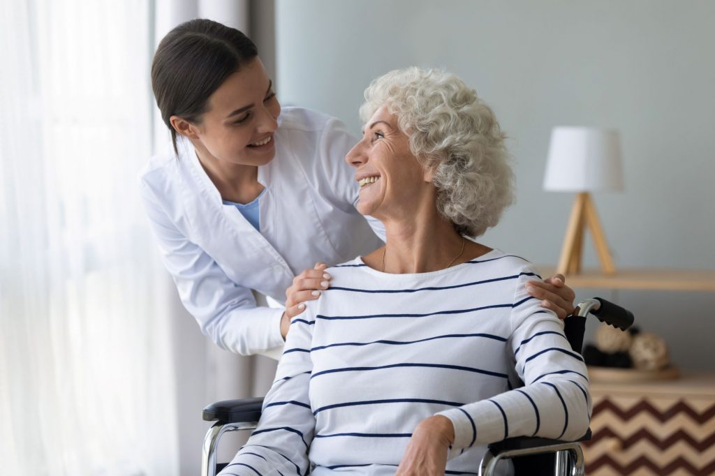 Older woman retiree with a white striped shirt, sitting in a wheelchair, smiling at a nurse who is helpng her 