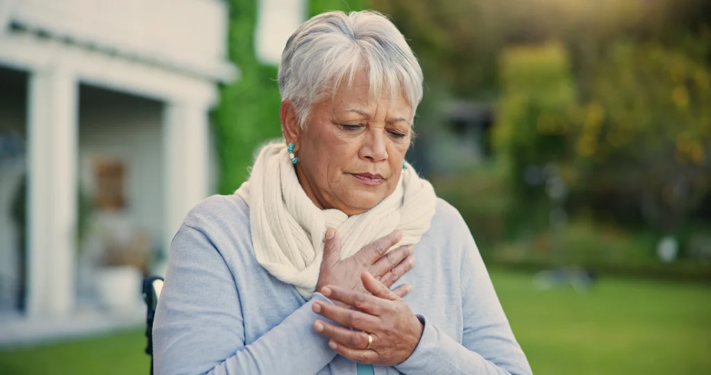 senior woman standing outside with hand over heart, maybe out of breath, maybe with chest pain