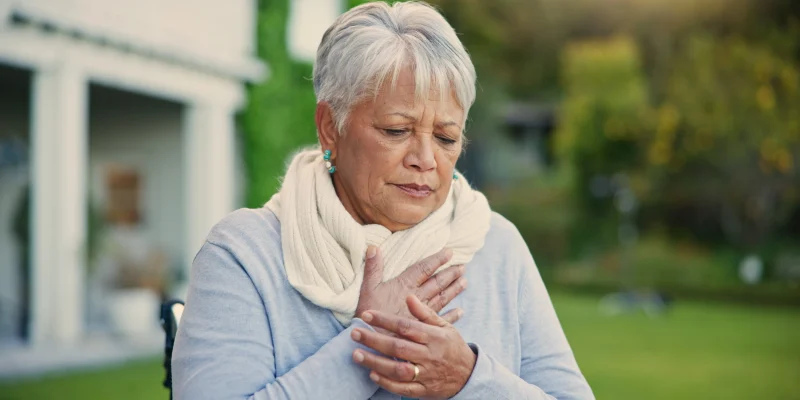 senior woman standing outside with hand over heart, maybe out of breath, maybe with chest pain