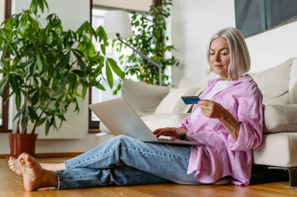 senior retired woman sitting on floor with computer and credit card in hand shopping online