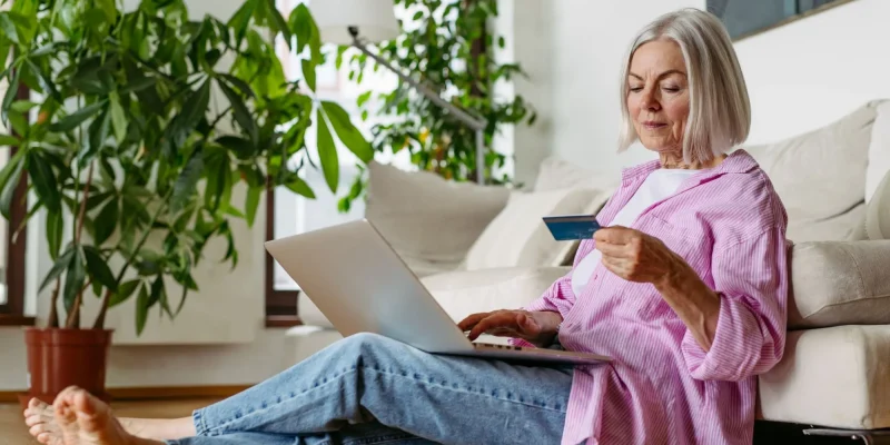 senior retired woman sitting on floor with computer and credit card in hand shopping online