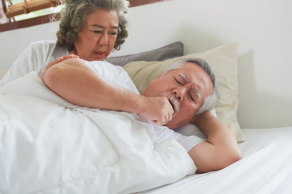 senior asian man in bed coughing with concerned wife looking over shoulder