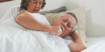 senior asian man in bed coughing with concerned wife looking over shoulder