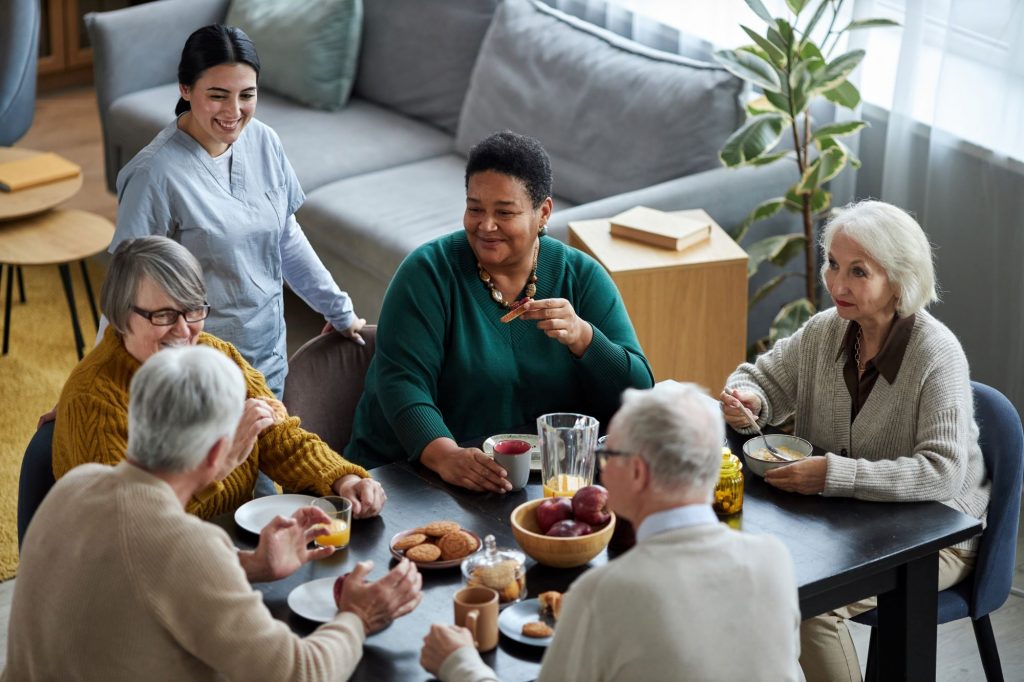 diverse group of older adults enjoying lunch with smiling female nurse in an assisted living community, probably in Seattle, Washington