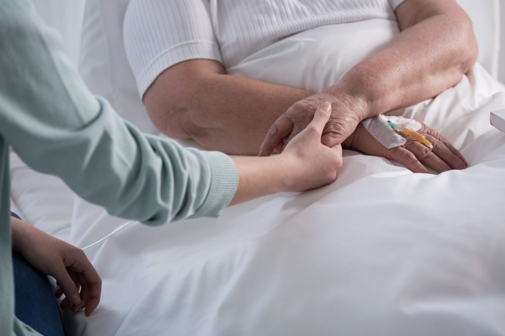 senior adult lying in hospital bed with iv in right hand and younger family member holding hand