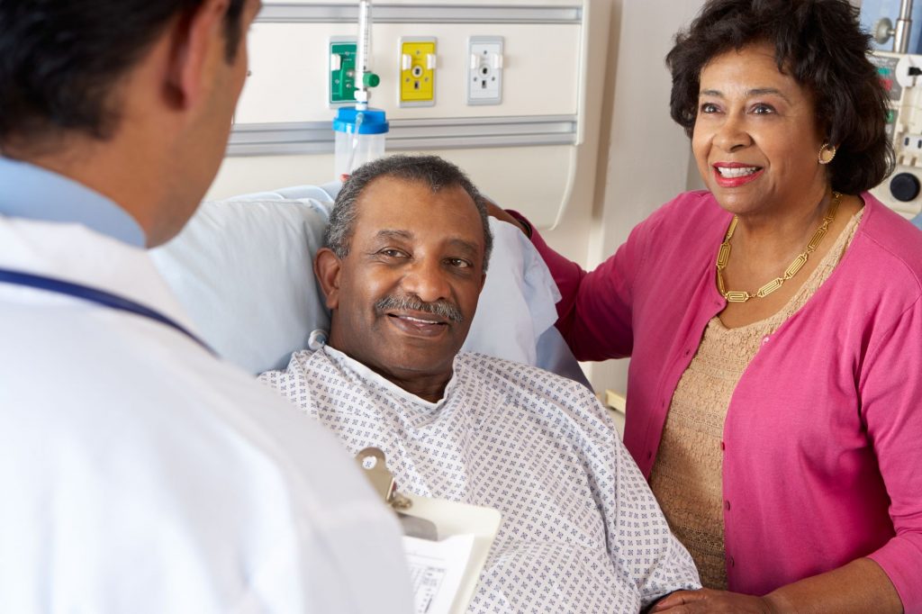 What You Need to Know About Medicare Spouse Coverage cover photo with a senior black man in a hospital bed with his wife by his side talking with a doctor