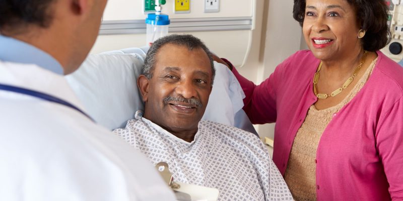 Black senior man in hospital bed with wife by his side talking with a doctor