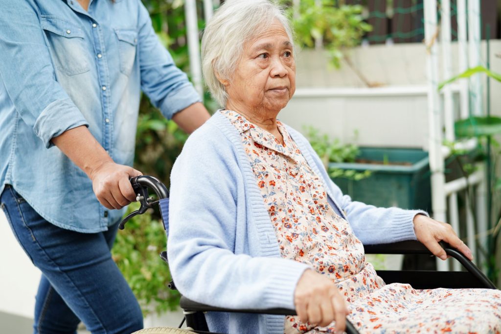 Elderly Asain woman being pushed in a wheelchair by a caregiver outside