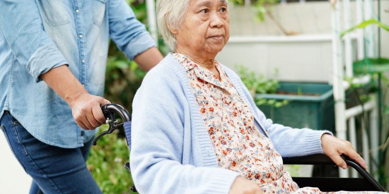 Elderly Asain woman being pushed in a wheelchair by a caregiver outside