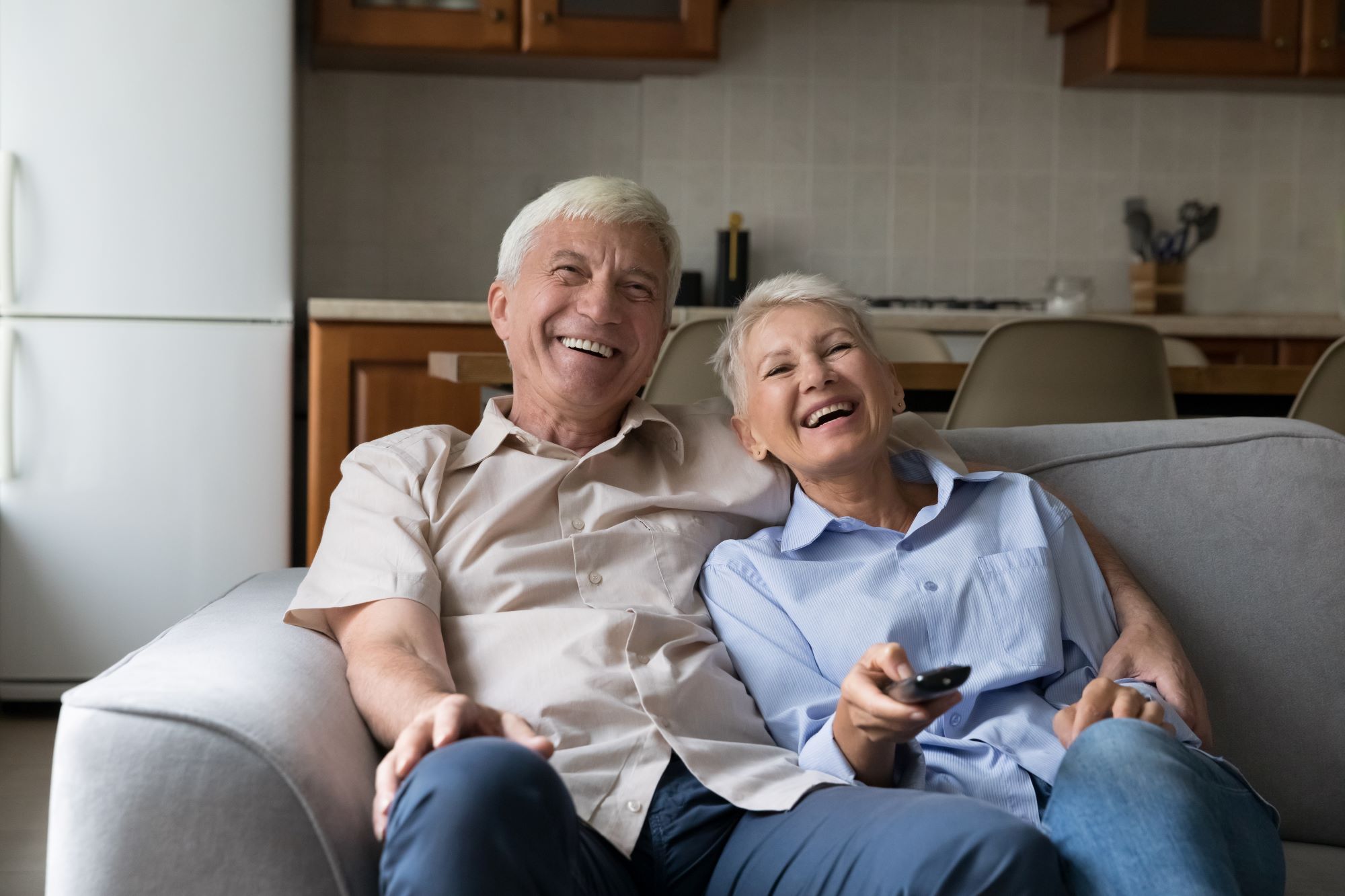 Laughing older couple relax on sofa spend carefree weekend together at home, hold remote control switch channels enjoy movie, watch favourite funny TV show use online stream services. Leisure concept