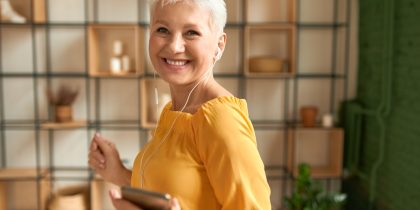 happy senior woman with ear buds smiling and holding phone, wearing a yellow shirt, short gray hair