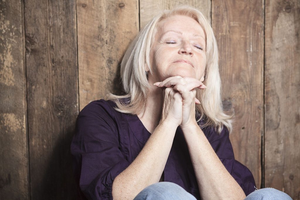 praying senior/retired woman with gray hair sitting against wood background with eyes closed in prayer