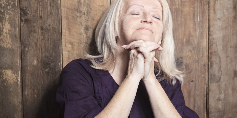 praying senior/retired woman with gray hair sitting against wood background with eyes closed in prayer