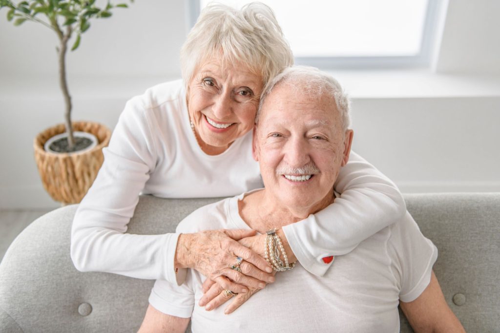 smiling seniors wearing white shirts, one sitting on the couch, the other hugging.