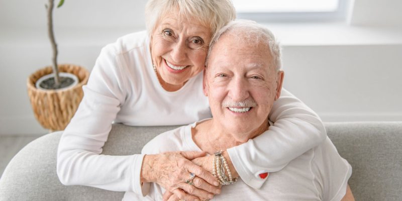 smiling seniors wearing white shirts, one sitting on the couch, the other hugging.