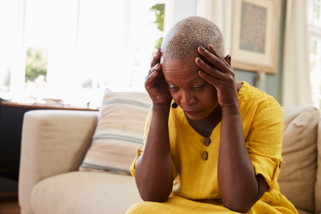stressed senior woman wearing a yellow dress and sitting on a couch with head in hands