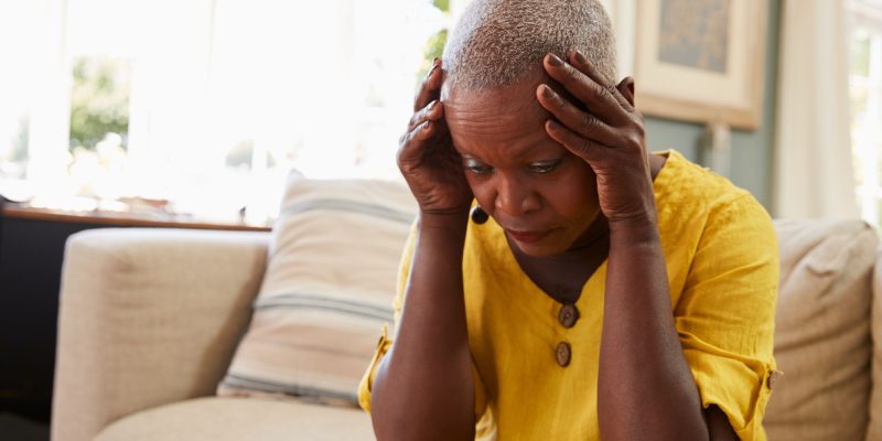 stressed senior woman wearing a yellow dress and sitting on a couch with head in hands