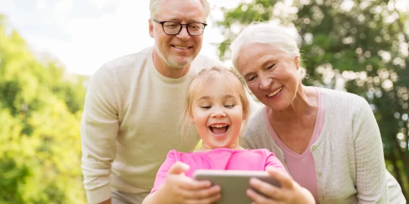 family, generation and people concept - happy smiling grandmother, grandfather and little granddaughter taking selfie by smartphone at park