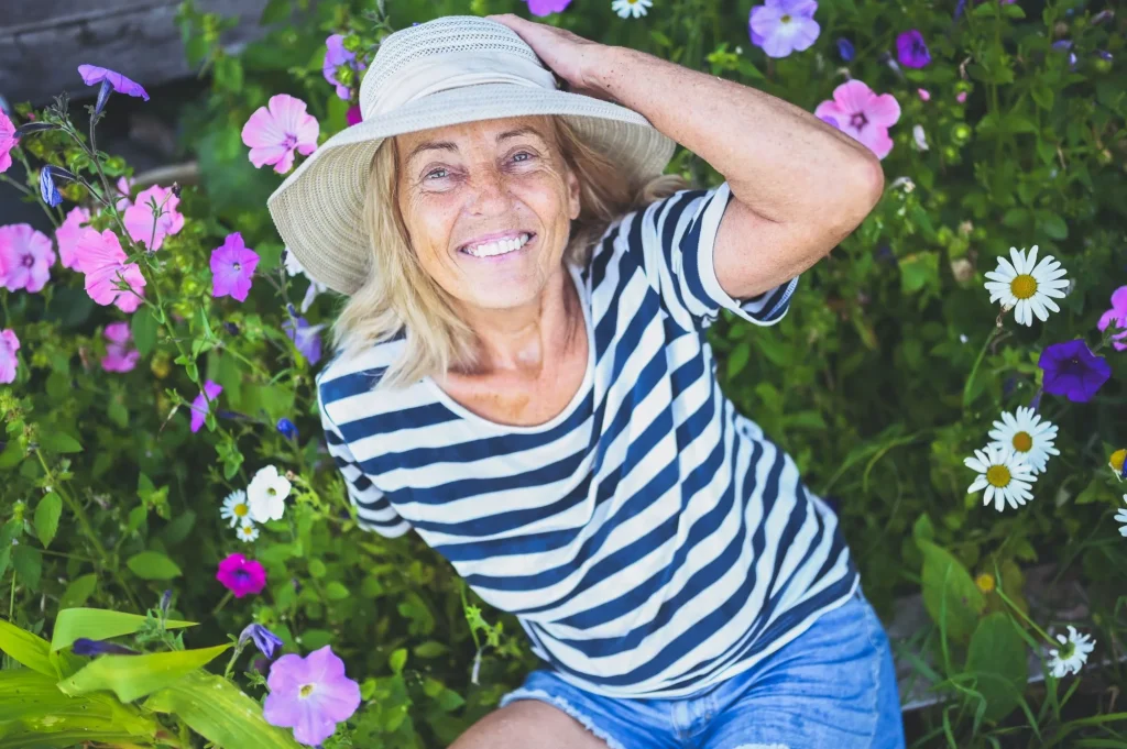 Happy smiling elderly senior woman having fun posing in summer garden with flowers in straw hat. Farming, gardening, agriculture, retired old age people concept. Growing organic plants on farm.