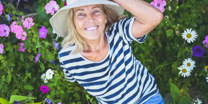 Happy smiling elderly senior woman having fun posing in summer garden with flowers in straw hat. Farming, gardening, agriculture, retired old age people concept. Growing organic plants on farm.