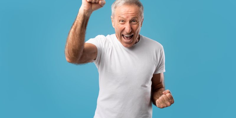 Overjoyed senior man screaming in excitement, gesturing yes shaking clenched fists on blue studio background. Triumphant elderly male celebrating his success, cheering favorite team at competition